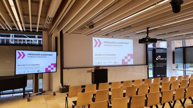 The picture shows an empty conference room. On the left hand side there is a TV screen showing the first slide of a powerpoint presentation with the title of the conference and the RHET AI logo. A second screen in the middle shows the same slide. On the right hand side you can see a roll up showing the RHET AI logo.