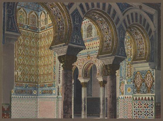 The image depicts a richly decorated architectural interior reminiscent of Islamic or Moorish design, specifically from the Alcazar in Seville, Spain. The space is characterized by ornate arches and columns that frame an intricate mosaic tile work on the walls.

Several large arched openings with detailed geometric patterns are visible, featuring a combination of vibrant colors such as blue, red, yellow, and green against white backgrounds. These tiles form complex medallions surrounded by smaller decorative borders within each alcove. The arches themselves have alternating dark and light stripes, adding to the overall ornate appearance.

The columns supporting these structures appear weathered or aged, suggesting historical significance. Some areas of wear are evident on the stone surfaces, which adds a sense of antiquity to the scene.

In the background, more intricate tile work can be seen in different sections along with additional architectural elements like pilasters and cornices that contribute to an overall impression of opulence and grandeur. The craftsmanship displayed indicates this may have been part of a palace or significant building designed for ceremonial purposes during historical times.
