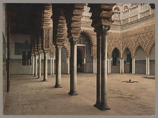 The image displays a traditional courtyard adorned with ornate arches and columns. The architecture showcases intricate designs, indicative of Middle Eastern or Moorish influences commonly seen in historical buildings such as Alcázar in Seville, Spain. The floors are composed of large stone tiles arranged in geometric patterns.

In the center stands a solitary lamp post on an open area surrounded by these architectural features. A few scattered objects can be observed: a small vase lies near one column and there seems to be another item placed further back against the wall or floor. 

The overall atmosphere is serene, with natural light filtering through openings above, casting shadows across the courtyard's surface. The image carries an antique feel, suggesting it may have been captured around late 19th century, potentially using a technique like photogravure as indicated by the descriptive information provided.