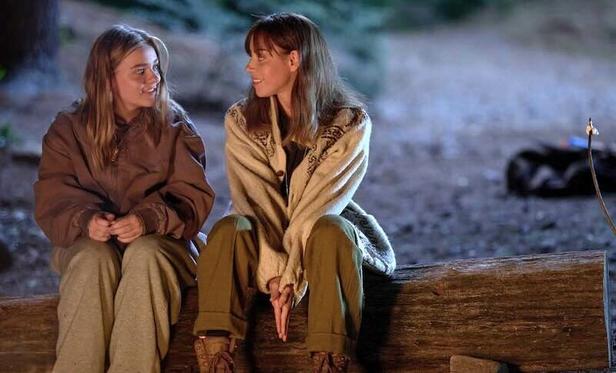 Maisy Stella and Aubrey Plaza sit on a log beside a campfire. They are looking at each other with smiles.