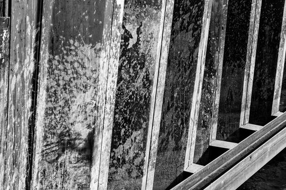 A black and white photograph of steel retaining supports. Looking like a fence or wall, it consists of alternating lighter and darker posts caused by light changes lining the left, with the rusted steel showing various kinds of texture and staining. To the lower right, just above a small bit of visible water, a steel i-beam from the support structure runs in parallel to the steel posts.