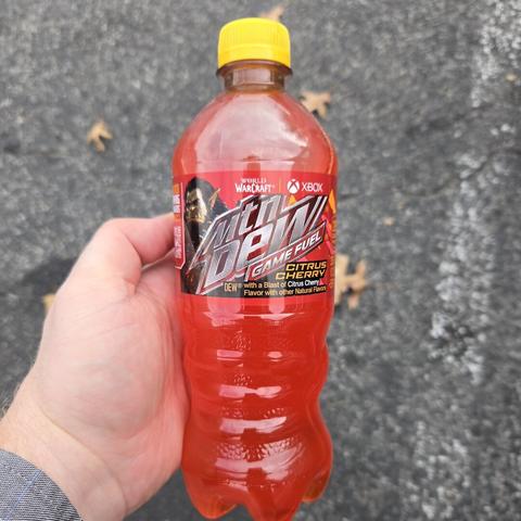 Photograph of a white person's hand holding a bottle of a reddish liquid (Mountain Dew Game Fuel Citrus Cherry) over an asphalt ground surface with some scattered leaves. The bottle is clear with a yellow cap and behind the silver Mountain Dew logo (stylized Mtn Dew), is an Orc from World of Warcraft with long, lustrous black hair.