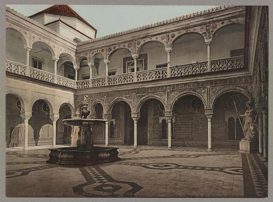 This image depicts an elegant courtyard with a central fountain as its focal point. The architecture is characterized by detailed masonry work, including ornate arches and intricate patterns on the walls. Balconies overlook this tranquil space from multiple levels of what appears to be a grand building or palace. Statues stand prominently within the setting, adding to the historical ambiance. The image has an old-fashioned tone with faded colors, indicative of a photograph taken many years ago.