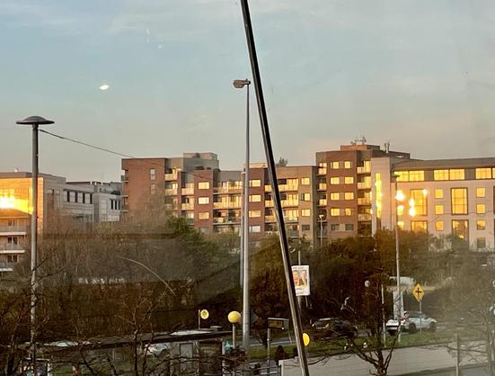 A cityscape at sunset, featuring modern buildings with glowing windows reflecting the light. Street lamps and trees are visible in the foreground, along with a sign on the side of the road.