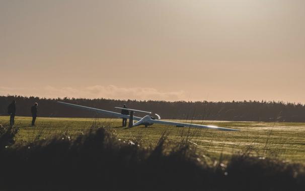A sunset shot of a glider waiting on a grass airstrip to take off. Several people are standing nearby.