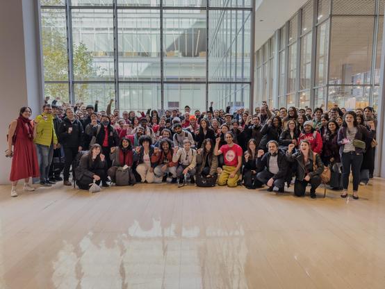 A group photo of about a hundred members of the Times Tech Guild, standing in the lobby of the New York Times building, many with fists raised in solidarity. In front of them is a shiny light wood floor, and behind are large floor to ceiling glass windows overlooking an inner greenery. Several members are wearing read dresses and shirts.