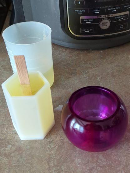 A white hexagonal-column silicone mold filled with molten yellowish wax, freezing to a white wax, with a flat wooden wick sticking out at top. Behind it a white silicone beaker with more wax (the leftovers). To its right, a purple epoxy plastic container or cup, squat and round, such as might be used as a planter or to hold such a candle. They are all sitting on a kitchen counter of brown and green mottled laminate.