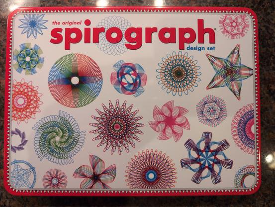 Photo of a spirograph toy/drawing tool in a metal tin. The tin is decorated with many, multicolored roulette geometry drawings.