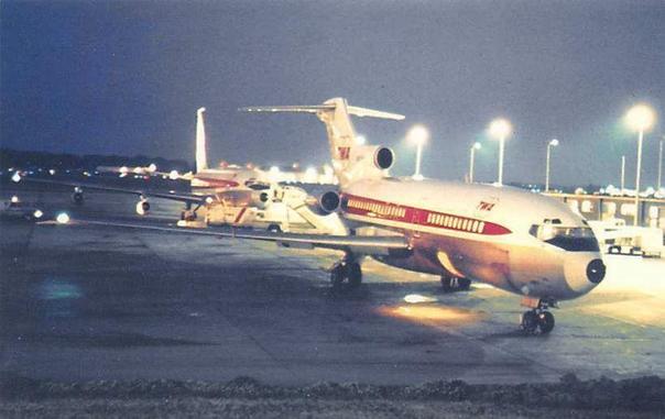 View of two TWA planes on the tarmac with lights on at night. Both are waiting for passengers next the terminal on thier right.