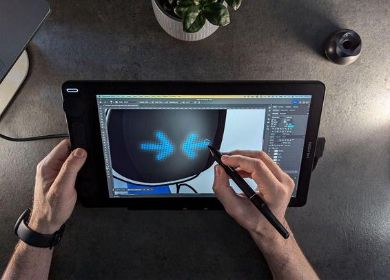 A person uses a stylus on a graphic tablet to edit a digital image of a robot. A small plant and a pen holder are nearby on the desk.