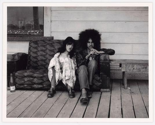 The image is a black and white photograph featuring two individuals sitting together outside on a wooden porch. The person on the left appears to be younger, with dark hair styled in an informal manner, wearing what looks like a patterned dress or blouse over casual pants. This individual has their legs crossed at the ankles.

On the right sits another figure who seems older and sports a voluminous afro hairstyle, dressed in darker clothing that suggests a leather jacket and trousers. They are holding onto something small with both hands on their lap while resting one arm casually across what appears to be an open suitcase beside them.

There's a patterned sofa or bench they're seated upon, which is detailed with dark fabric featuring decorative swirls or paisley designs against a lighter background. In front of the figures lies a glass bottle labeled 'trip,' indicating it might contain alcohol. The setting seems somewhat casual and lived-in, perhaps in a rural environment given some visible foliage through the window behind them.

The image has an overall candid quality to it with natural lighting that suggests daytime. There's also text or graffiti on what looks like a wall above the sofa which is not entirely legible due to resolution constraints but contributes to the informal atmosphere of the scene.