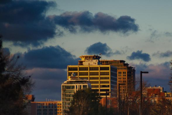 A photograph of several tall concrete buildings shot from below, increasing in height as they get further away. The one in the middle has a helipad atop it. Dark clouds loom in the sky, moving in from the left.