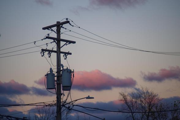 A photograph of a power pole with three transformers on it. Fluffy blue and pink clouds hover behind it.