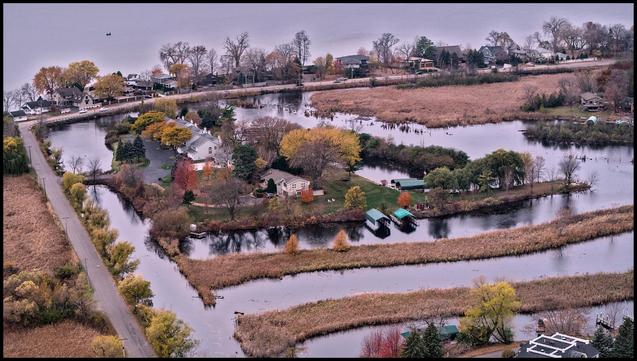 Ariel view of a human made harbor with channels and strips of dark brown swamp grass. Some homes are built on a island in middle of small harbor and a road borders harbor on top and left sides of image.