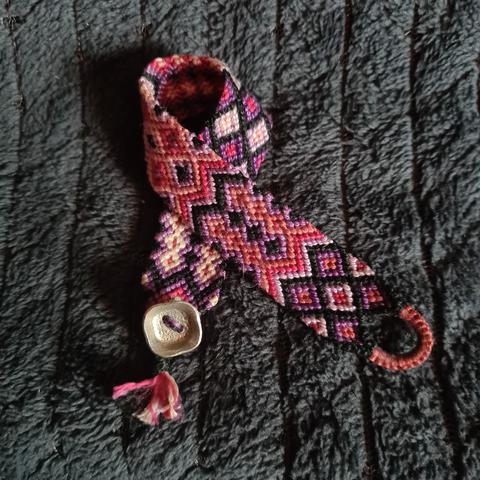 Un bracelet en macramé avec plein de formes géométriques dans les tons rose, parme, violet, noir, avec un bouton de métal argenté carré aux bords arrondis...