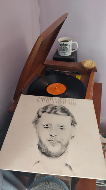 Record player with a 12" black vinyl record. In the foreground is an album cover showing Harry Nilsson's face, with the features doubled. On the corner of the player is a mince pie, and behind is a mug of coffee on a speaker.