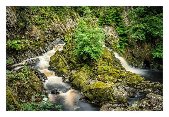 A small river divides around a rocky outcrop into two waterfalls. The rock is mossy and has a single tree growing at its summit.