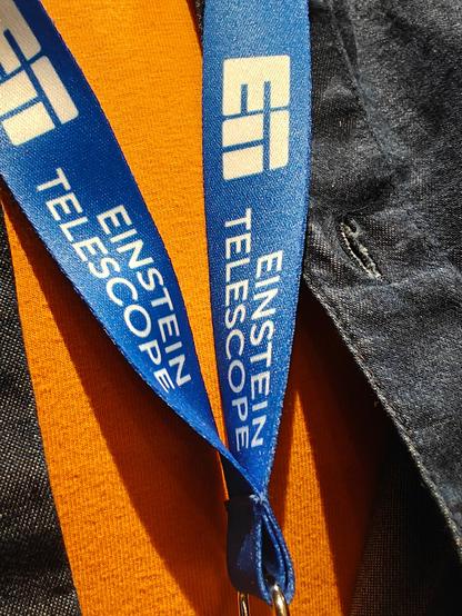 A close-up of a blue lanyard with (in white) the words 'Einstein Telescope' and the logo of ET. One can see the person with the lanyard is wearing an orange t-shirt and a dark blue shirt.