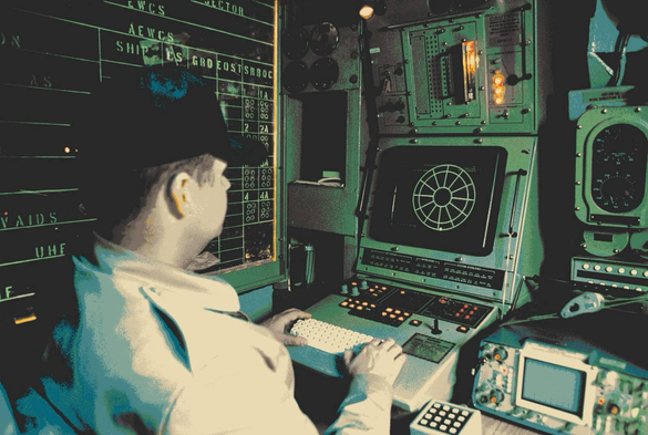 Some guy wearing a cap using a retro computer terminal probably on a submarine or military installation.