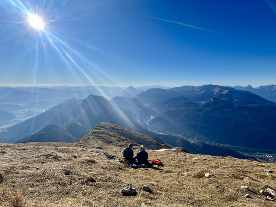 Bergpanorama mit 2 in der Wiese sitzenden Personen und Sonnenstrahlen direkt auf die 2