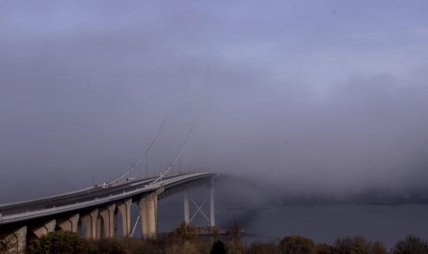 The Forth Road bridge partially obscured by fog with sunlight on Autumnal trees in the foreground