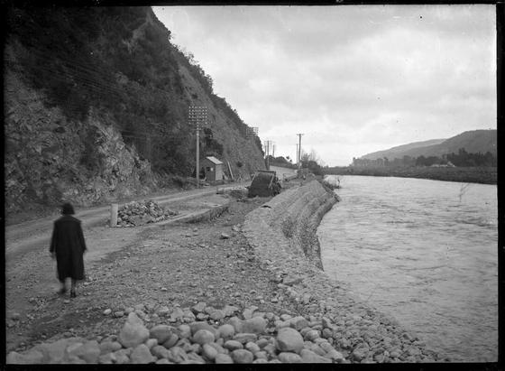 Black-and-white photo: By the Hutt River, Wellington, in the Taita Gorge area. 1910s-1930s? Photographer, A. P. Godber. Description: A view in the Taita Gorge area, above the Stokes Valley entrance, which includes a newly constructed embankment constructed of boulders bound with wire. Inside the wall a tip truck is filling the gap between the wall and a road. To the left of the road is a steep hillside. A small shed is visible in the centre of the image, and poles carrying power lines run along the left side of the road. Citation: Godber, Albert Percy, 1875-1949 :Collection of albums, prints and negatives. Ref: APG-2103-1/2-G. Alexander Turnbull Library, Wellington, New Zealand. https://natlib.govt.nz/records/23224015