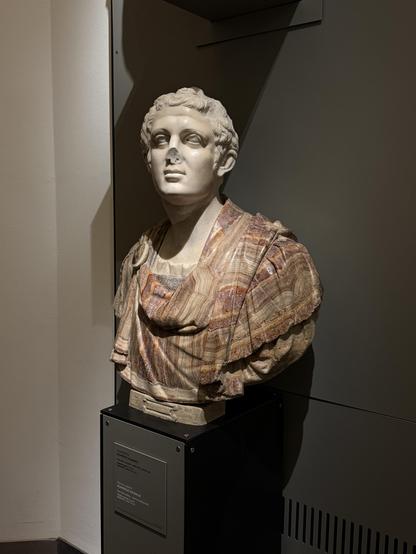 A statue of a head & bust on display at a museum, mounted on a plynth.  The chest is covered in a colourful stripey “material” that has lines of pale reds, yellows, beiges, brown and orange.  The sculpted clothing appears to drape on the body, with the stripes correctly following the shape as if the material were cloth, not marble.