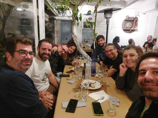Picture shows all members of the org team sitting in a rectangle table that has lots of food and drinks on it. Everyone is smiling and looking at the person taking the photo which is a selfie.