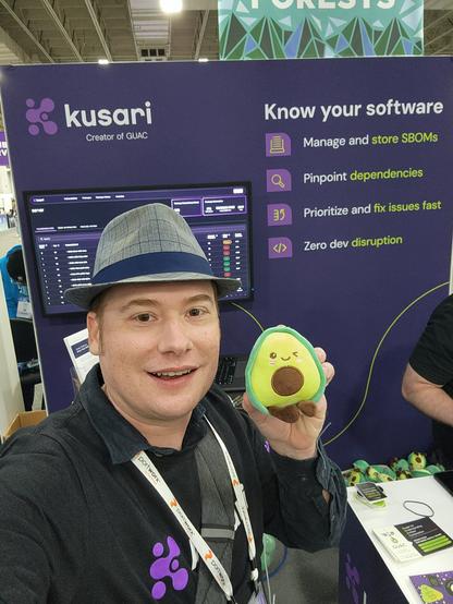 A silly looking man wearing a hat and holding an avocado plush.