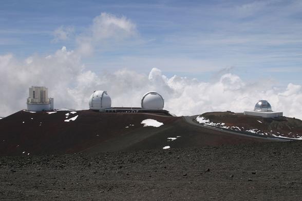 Four white and silver observatories lined up along a ridge on top of Mauna Kea. There are clouds at the same elevation in the background, and scattered snow pockets on the ground.