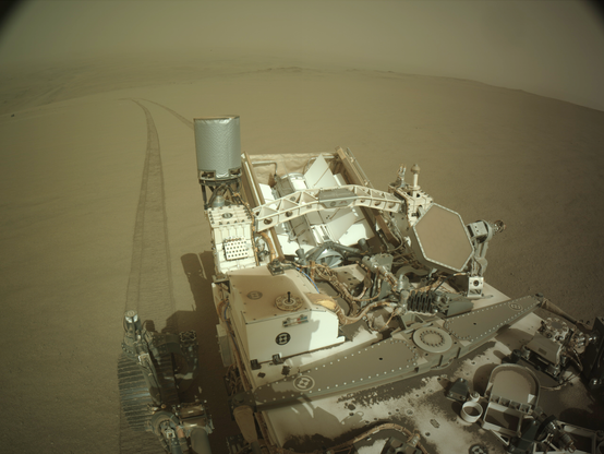 Le rover Perseverance est visible au premier plan. Derrière lui, des traces de roues se dessinent sur le sol lisse et se perdent peu à peu dans le paysage en formant une légère courbe. À l'arrière-plan, on aperçoit l'intérieur du cratère Jezero.