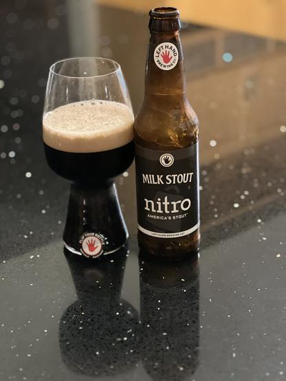 A stout glass of dark beer with a thick tan head. The brown bottle is to the right with a black label with white lettering. The neck label has the red Lefthand left hand logo. The bottle cap, also with a red hand, is balanced on the glass