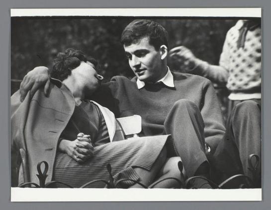 The image depicts a moment between two young individuals, presumably in an outdoor setting such as a park. The younger man with dark hair is wearing a sweater and appears to be seated on another person's lap or beside them. He has his arm around the shoulder of what seems to be a younger woman who rests her head against him, displaying affectionate body language.

The image captures this intimate moment in black-and-white, giving it an enduring quality that might suggest a scene from mid-20th century photography. The focus is on their expressions and physical connection rather than any background details, which are intentionally blurred or indistinct to emphasize the subjects' interaction.

This photograph may have been taken by Ed van der Elsken, as indicated in the provided source information. Van der Elsken was a Dutch photographer known for his work documenting everyday life across Europe during various decades. The image is likely part of his body of work captured between 1950 and 1954.