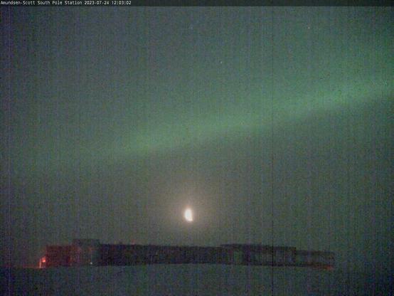 A grainy night-time webcam picture of a research station at the South Pole, a large building low to the ground.

Above it is a bright green light, across the night sky, and right between it and the research building is a brightly lit up crescent moon.