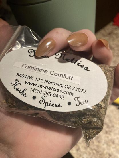 A hand holding a small package labeled "Feminine Comfort," containing a blend of herbs and spices, with a website and contact information printed on the label. The background shows a blurred countertop.