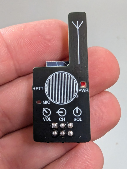 A tiny circuit board in the shape of a hand held radio, held in a hand.