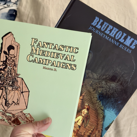 A hand holding the book covers of Fantastic Medieval Campaigns and Blueholme Journeymann Rules.
