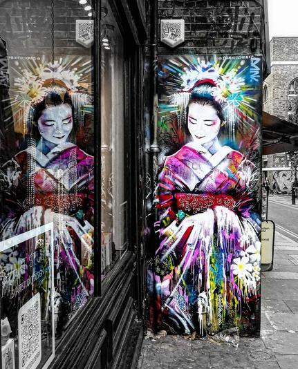 Streetartwall. A beautiful mural of a colorful geisha was sprayed on a small black brick wall next to a store. The beautiful Asian woman with a white-painted face holds her arms out in front of her in a kimono, surrounded by flowers and an explosion of graffiti colors. The sophisticated photo is taken in such a way that the geisha is practically looking at herself in the mirror of the shop window.