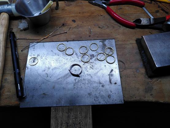 Photo of 8 brass rings and one silver ring placed on a small metal surface integrated into a wooden workbench. Pliers, hammers and a bucket of flux appears in the periphery.