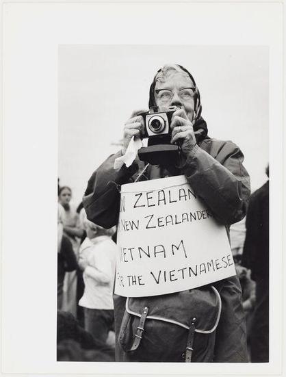 The image depicts a person holding up an old-fashioned camera to take a photograph. They are wearing glasses and have grey hair, with some red paint or ink on their face around the eyes area, suggesting they may be part of a protest movement. The individual is also dressed in dark clothing, including what appears to be a coat or jacket. Around their neck is draped another piece of clothing that looks like it might be protective gear for protests.

In both hands, the person holds up signs with text on them. One sign reads "NEW ZEALANDER NEW ZEALANDERS VIETNAM" while the other says "VIETNAMESE." These messages indicate a connection to Vietnam and its Vietnamese population or citizens. The context of this image seems related to protests, possibly against American military involvement in Vietnam.

The background is not entirely clear but suggests an outdoor setting with some people standing around, hinting at a gathering for either the photograph being taken or another event taking place simultaneously.