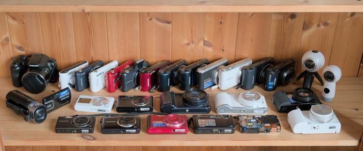 A wooden shelf with three rows of cameras, top to bottom, left to right:

WB1100F, WB31F, 3x WB37F, WB50F, WB150F, WB200F, WB250F, WB351F, WB380F, WB800F, WB850F, Gear 360 from 2017 and from 2016

HMX-QF30 camcorder, DV150F, DV300F, Galaxy Camera, Galaxy Camera 2, EX2F

SH100, ST150F, ST200F, ST1000, ST1000 disassembled, EX2F