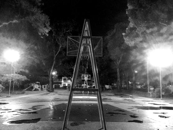 an empty basketball court in a park at night seen from the far end, looking through one "A" shaped goal post to the other. Bright lamps on the left and right light up the night with trees overhead.