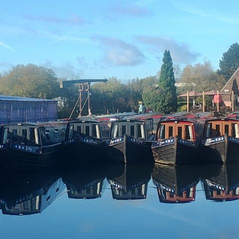 A row of hire boats floating in a basin that's perfectly still. The boats are seen head on. Their bows are reflected in the water.
Behind them is a manual lift bridge and some trees. The sky is light blue with some clouds. It's reflection in the water is even bluer.