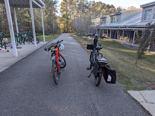 Photo of two bicycles, side-by-side, waiting for their riders on a greenway outside an elementary school. To the left of the greenway is a covered bike parking area; to the right is the school. Just out of frame is a dedicated school entrance for kids who show up on foot, bike, or scooter.