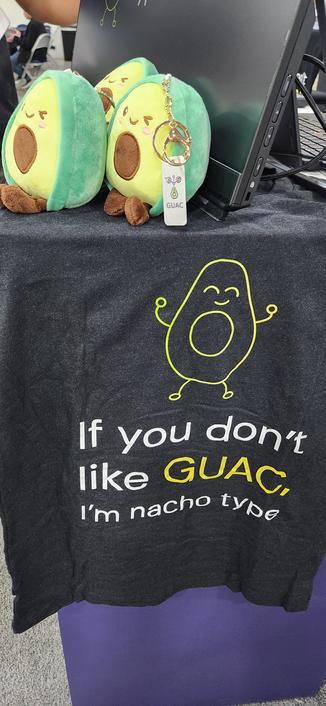 A t-shirt that says "if you don't like GUAC, I'm nacho type" hanging off the side of a booth desk. Stuffed avocado plushies sit on the top.