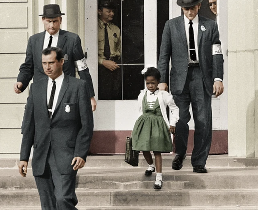 Iconic photograph of 6yo Ruby Bridges, beautifully groomed and dressed with pride by good, loving parents, being evicted on her first day and escorted by Federal Marshalls away from her all-White elementary School. The pain and suffering from the humiliation and trauma are evident in her face.