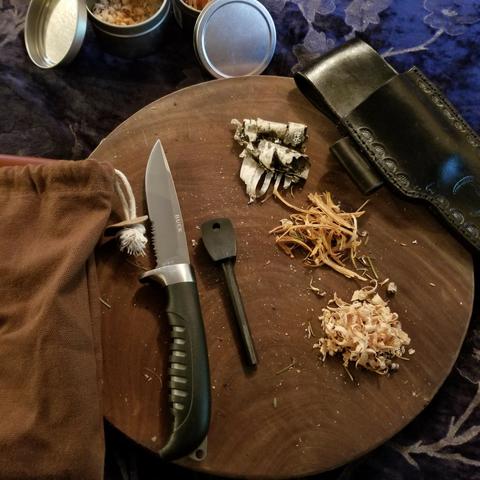 A fire starting kit with tinder materials (birch bark, fatwood slivers and fatwood curls), a ferro rod, a Buck knife with a small section of serrated edge for making the slivers and curls, a cotton pouch, a black leather knife sheath with a side loop for the ferro rod. Small tins to hold the tinder. All the items are sitting on a wood cutting board.