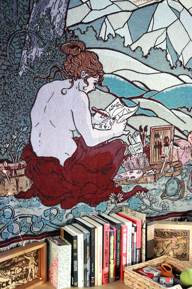 a closeup of the tapestry showing the figure leaning over to pain. It is inside hanging on a walls and there are books and boxes beneath it.