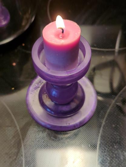A burning short violet cylindrical candle in a violet plastic candlestick, on a glass radiative stovetop.