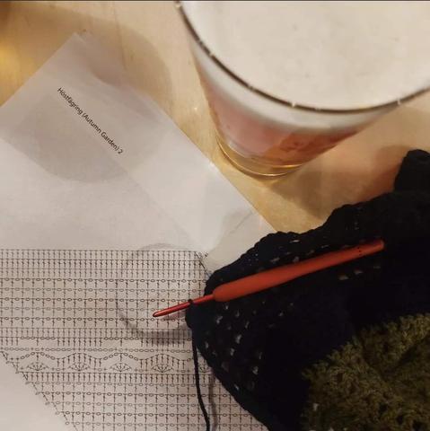 Pub table viewed from above: a crochet pattern, hook with a scarf in the making and a glass of Dortmunder
